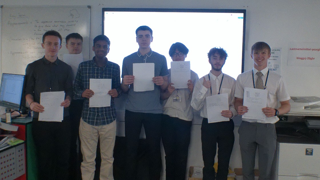 A group of students standing in a classroom holding up papers, with a whiteboard in the background