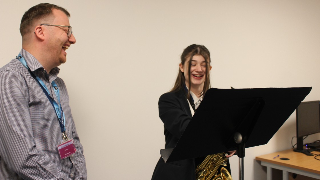 Teacher and student smiling during a music lesson, with the student holding a saxophone and standing at a music stand