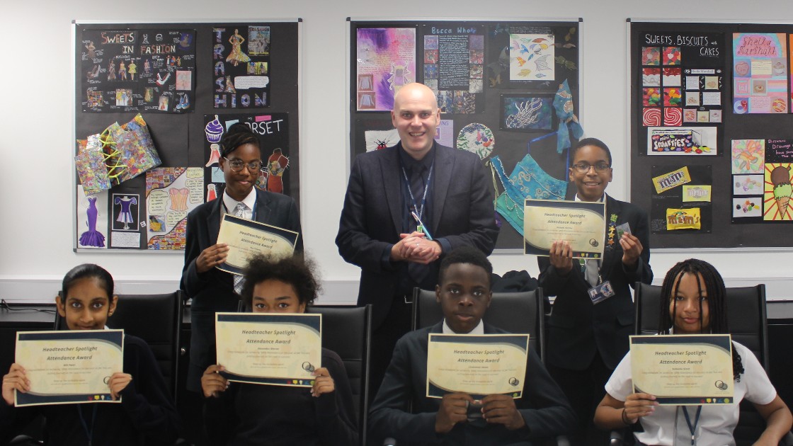 A headteacher stands with six students holding certificates in a classroom, with student artwork displayed on the wall behind them