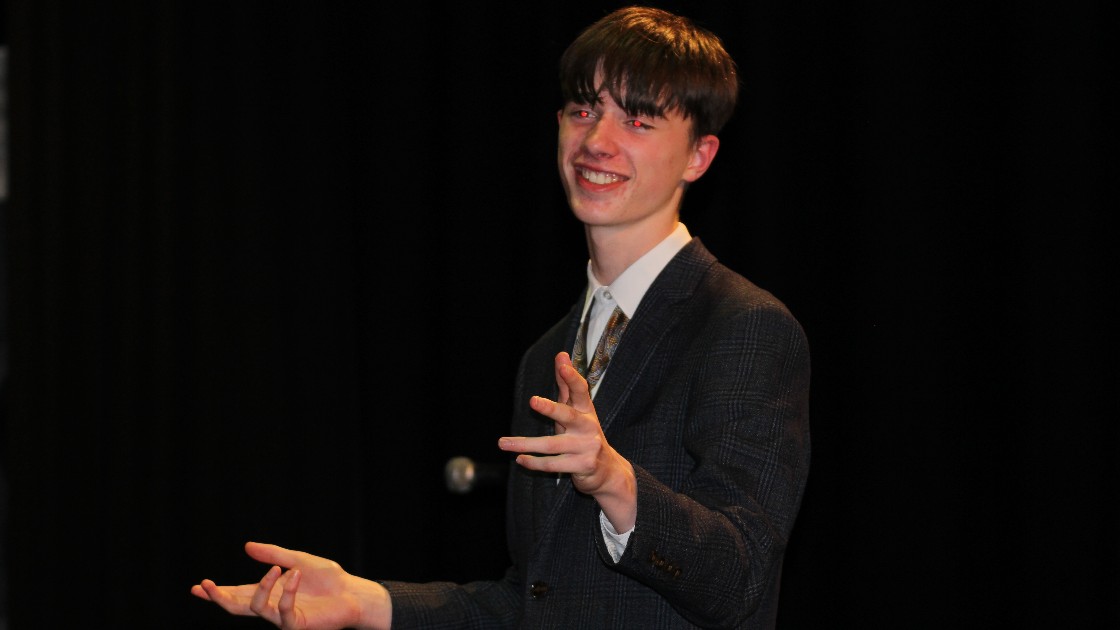 Student in a suit smiling and gesturing on stage performing in Summer Fest Day One