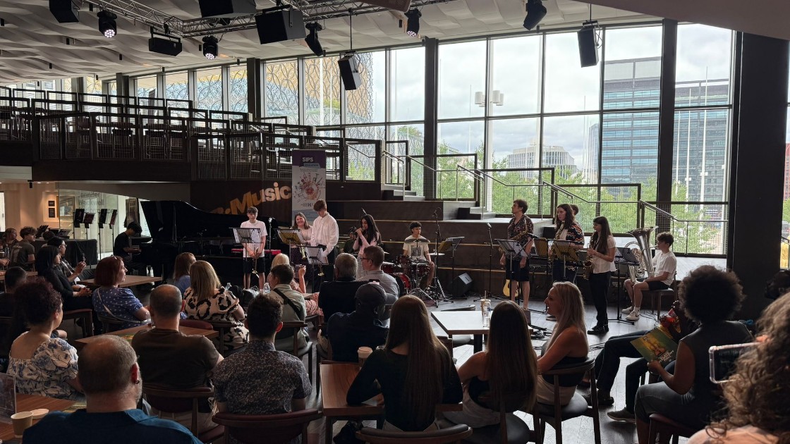 A student band performing on stage in front of an audience in a modern venue with large windows