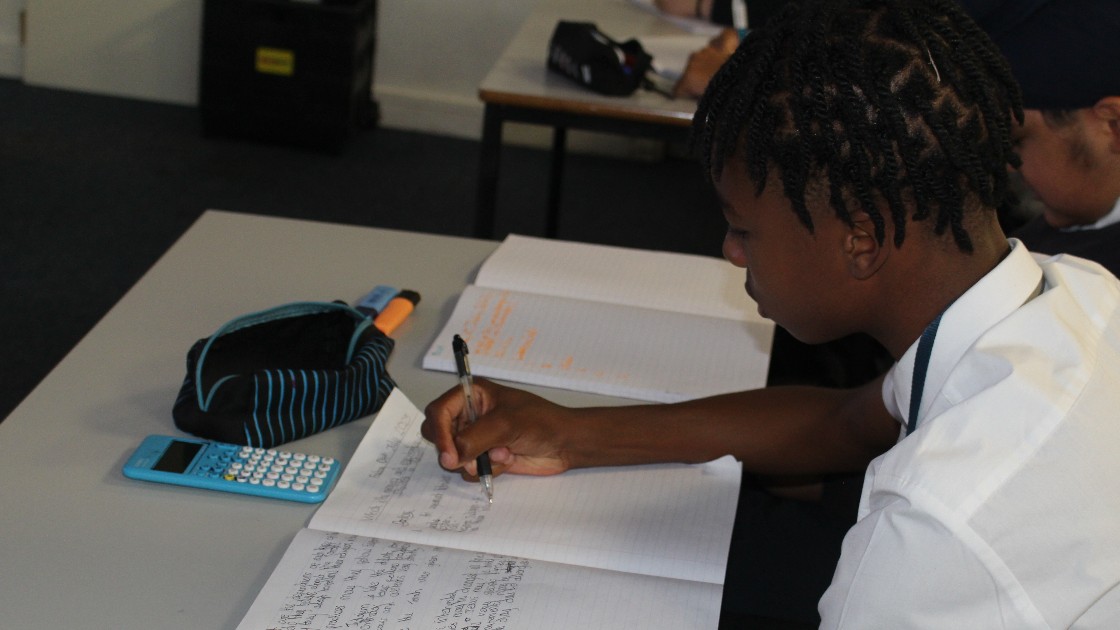 Student writing in exercise book during a lesson, with a calculator and pencil case on the desk