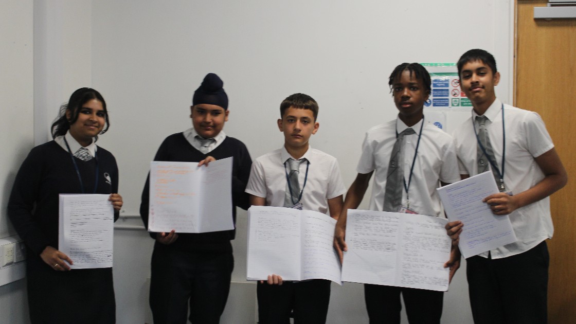 Five students in uniform holding open exercise books, standing in a classroom