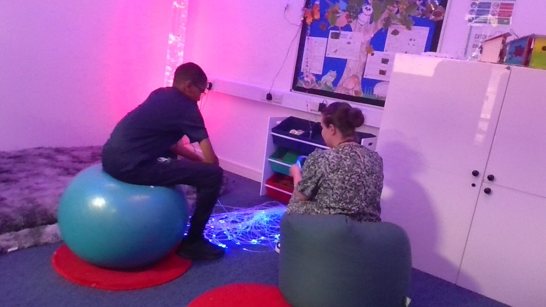A student and a teaching assistant sitting on exercise balls in a sensory room with glowing fibre optic lights